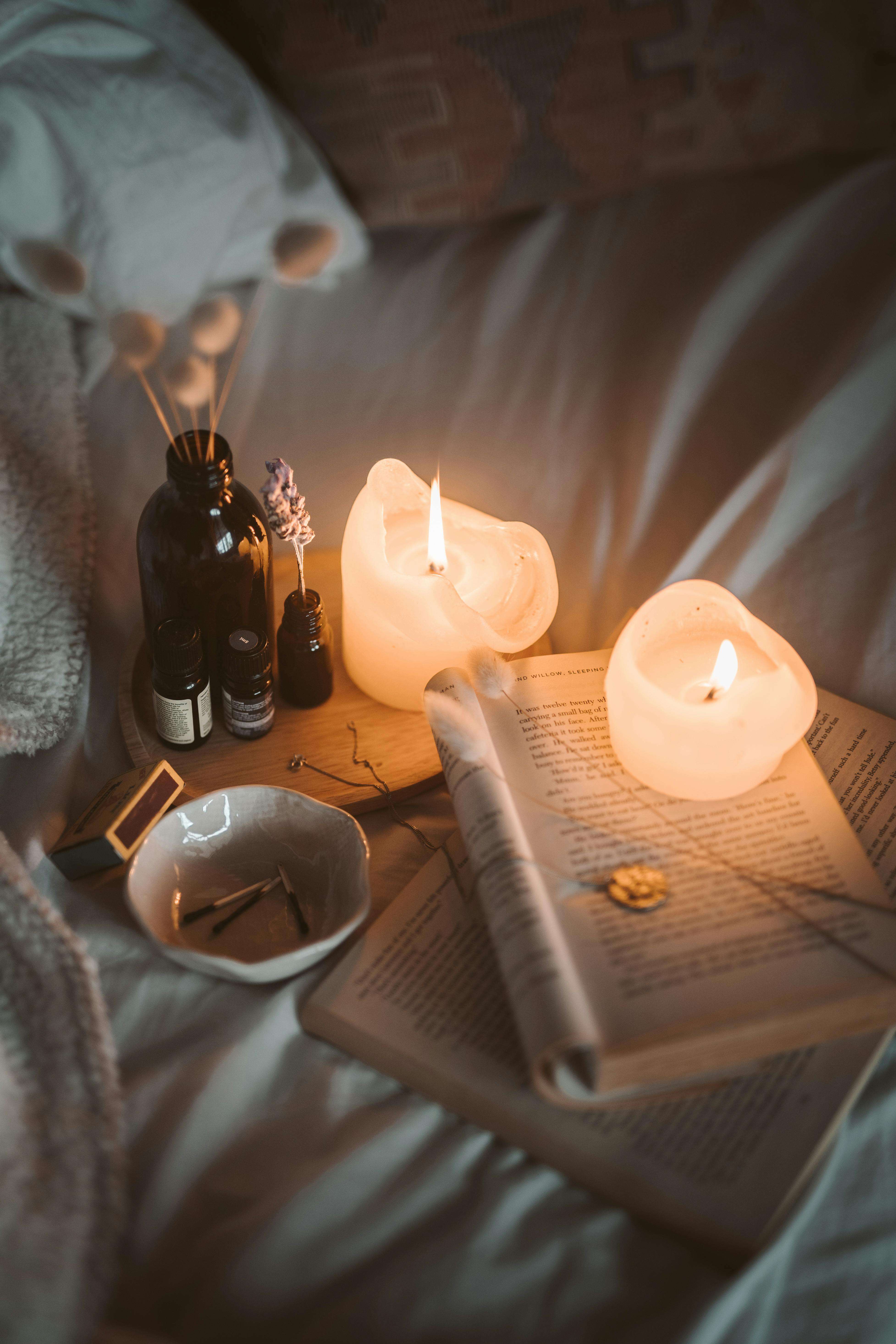Cozy spa-like table with lit candles, essential oil bottles, dried lavender, and an open book on a wooden surface.